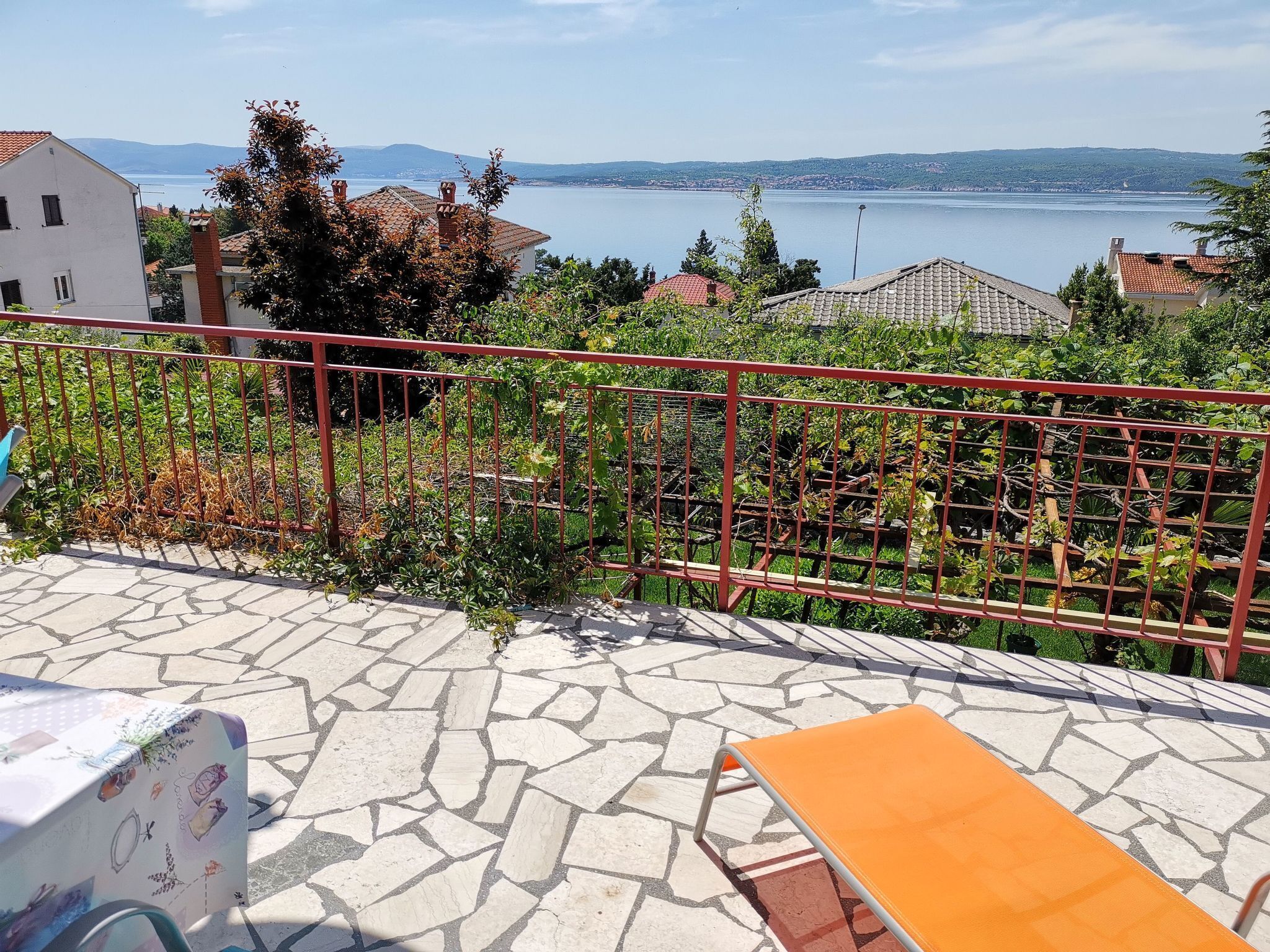A terrace with a stone floor, a red metal railing, and a view of a lake and distant hills.