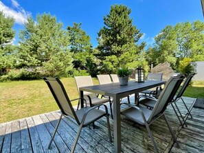 Eine Terrasse im Freien mit einem rechteckigen Tisch und sechs Stühlen, umgeben von einem Holzboden und einer Rasenfläche mit Bäumen.