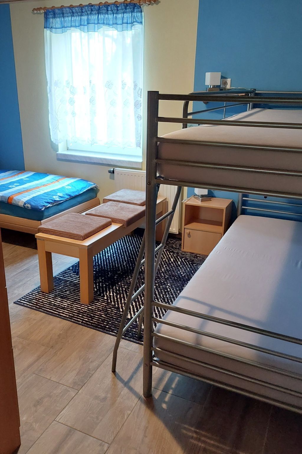 A bunk bed with a wooden bench and a small table, a window with a floral curtain, and a blue wall.