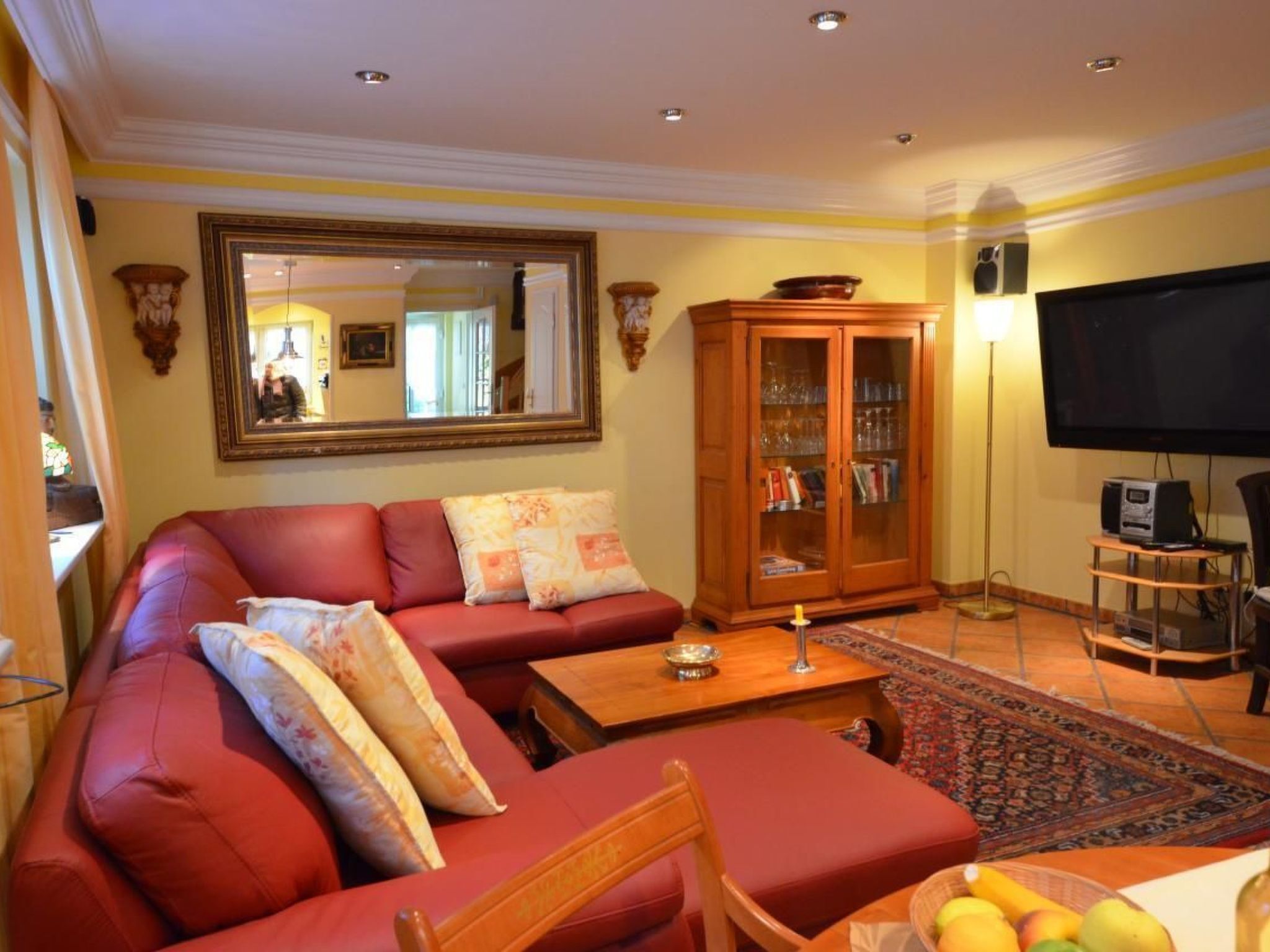 A living room with a red sofa, a wooden cabinet with glass doors, a flat-screen TV, and a dining area with a table and chairs.