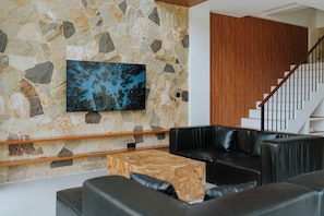 Uma sala de estar moderna com uma parede de pedra, uma TV de tela plana, uma mesa de centro de madeira, sofás de couro preto e uma escada de madeira.