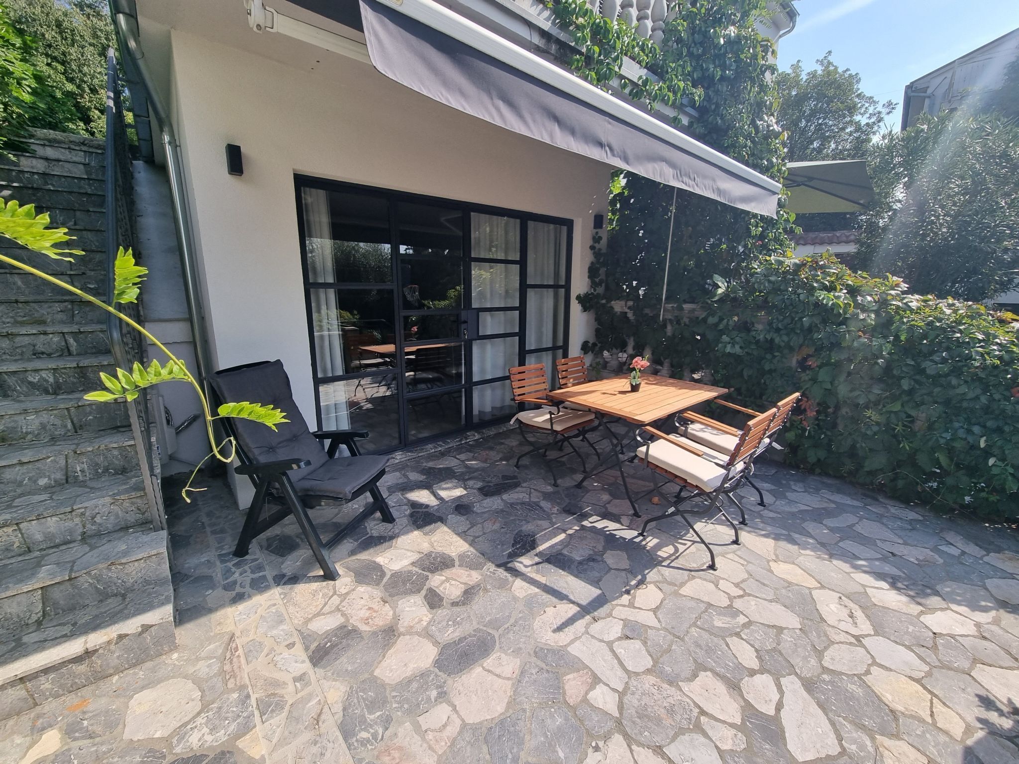 En terrasse med stengulv, et træbord med stole og en glasdør, der fører til et indendørs område.