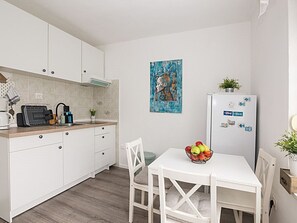 A compact kitchen with white cabinets, a wooden countertop, a white dining table with a bowl of fruit, and a refrigerator with magnets. - Charmante Wohnung in Crikvenica für 2 Personen by Interhome (Crikvenica)