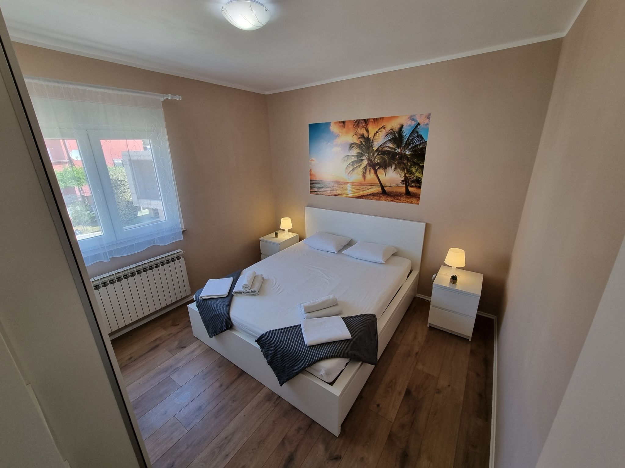 A bedroom with a large bed, two bedside tables with lamps, a window with a view of buildings, and a painting of a beach scene.