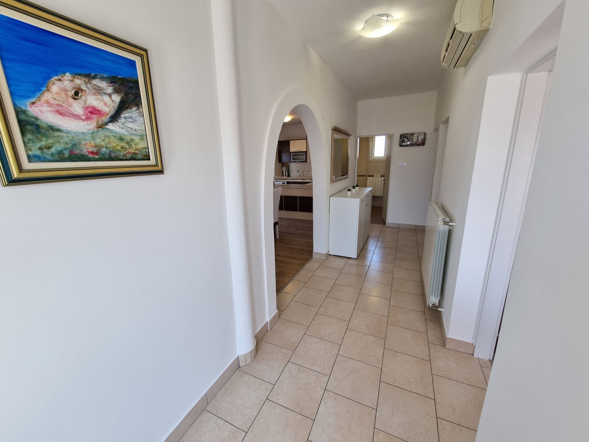 Un couloir avec un sol carrelé, un tableau encadré au mur et, au fond, un coin cuisine.