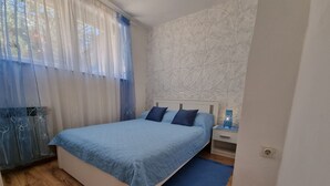 Un dormitorio con cama, mesita de noche, lámpara de pared y ventana con cortinas translúcidas.