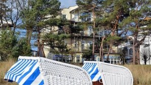 Una playa con dos sillas blancas de mimbre con fundas a rayas azules y blancas, arena y un edificio con varios balcones al fondo.