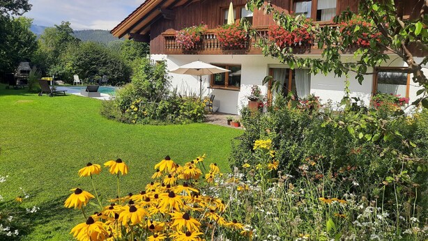 Una casa de madera de dos pisos con balcón, rodeada por un jardín cuidado con flores amarillas y una piscina.