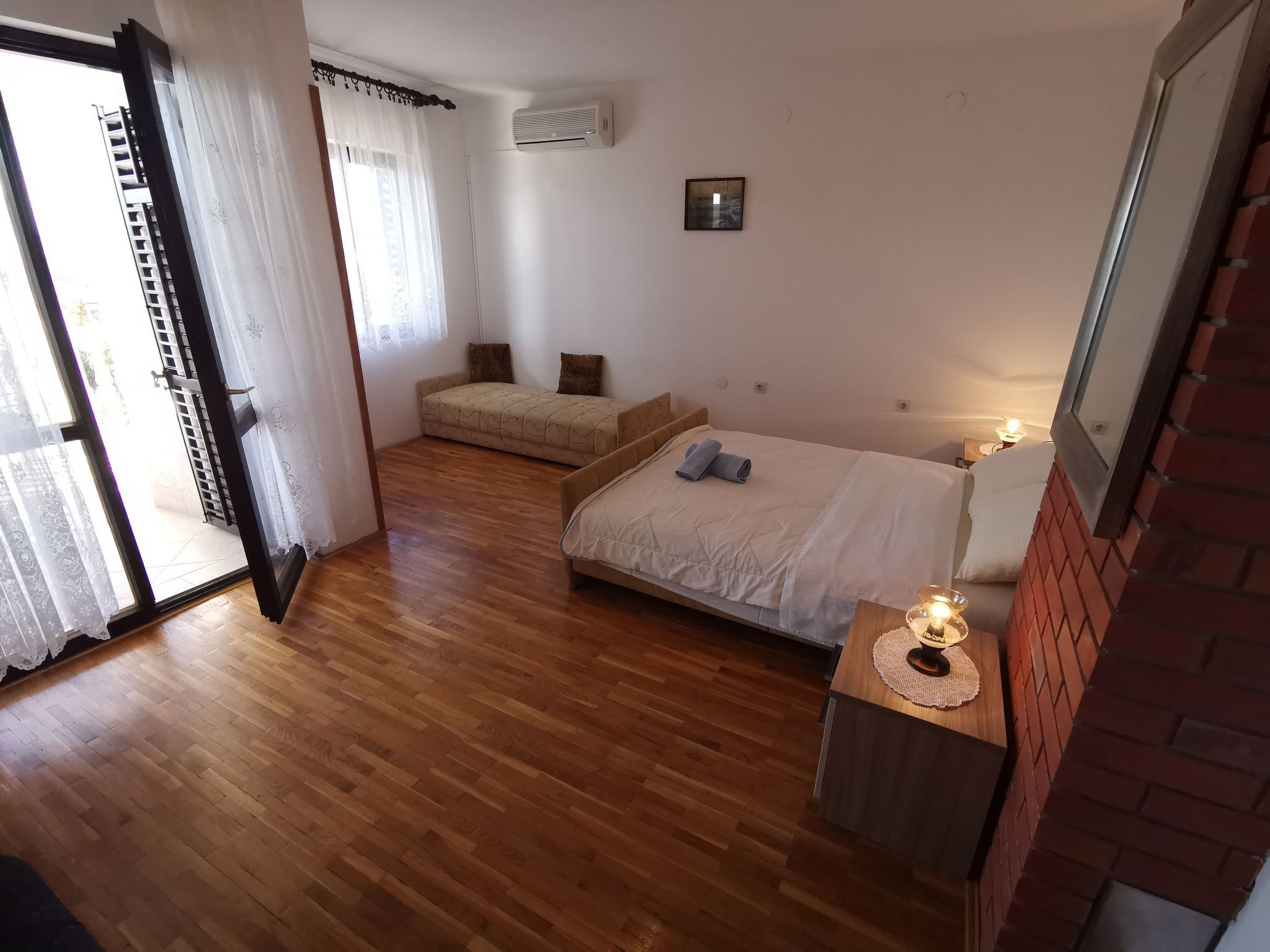 A bedroom with a bed, a small sofa, a bedside table with a lamp, and a sliding glass door leading to a balcony.