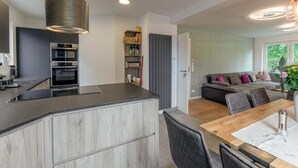 Una cocina moderna con electrodomésticos integrados, una zona de comedor con mesa y sillas de madera, y una sala de estar con sofá y cojines.