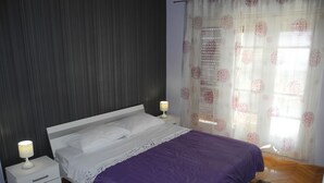 Un dormitorio con una cama, mesitas de noche, una lámpara, un tapete estampado y una ventana con cortinas traslúcidas.