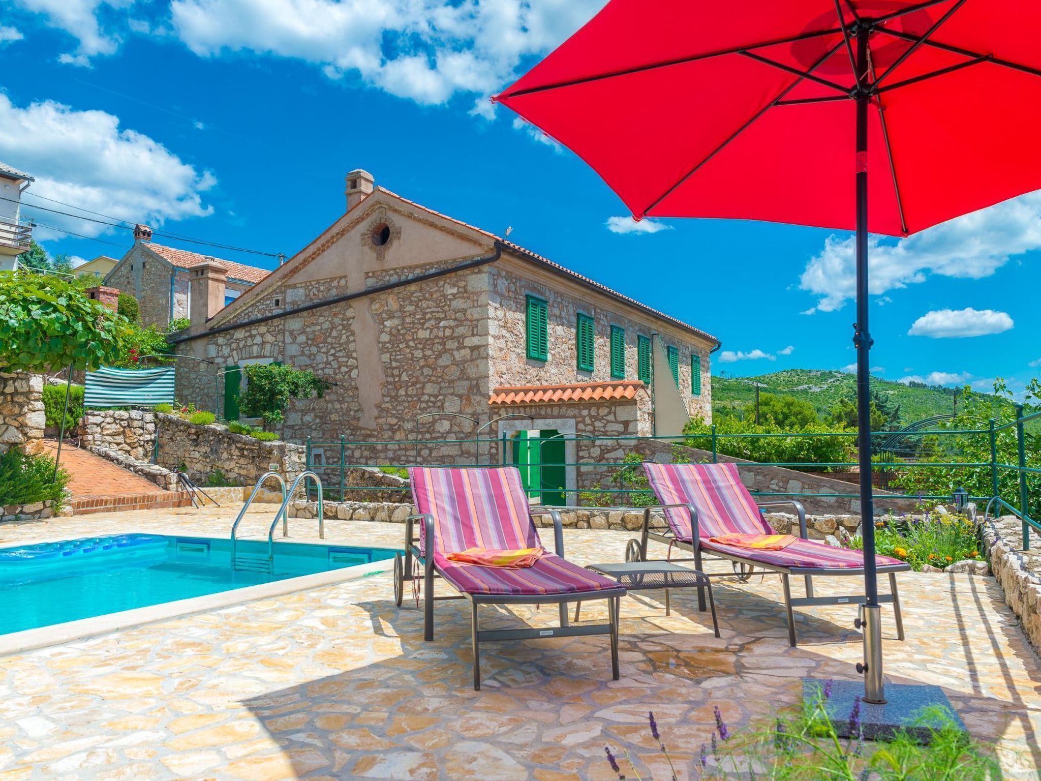 A stone building with a pool, two lounge chairs, and a red umbrella.