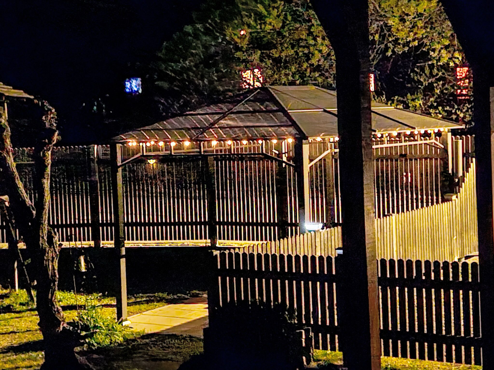 A gazebo with a metal roof and string lights, surrounded by a wooden fence.