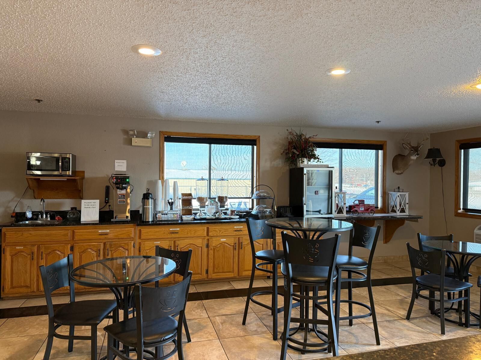 A modern kitchen with wooden cabinets, a microwave, and a coffee station. There are several chairs and a glass table.