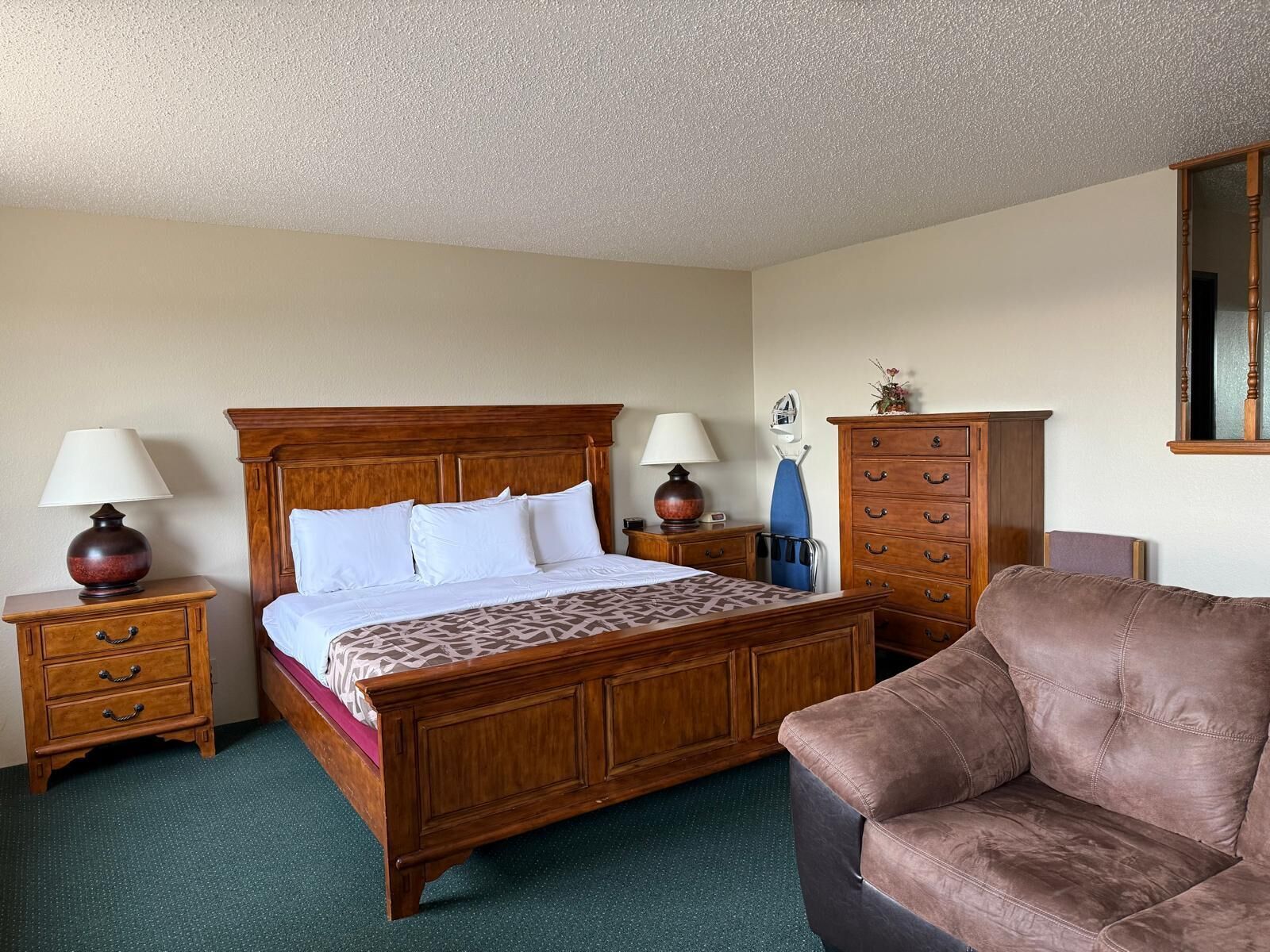 Une chambre d’hôtel avec un lit en bois, deux tables de chevet avec des lampes, une commode en bois, un canapé en cuir marron et un miroir au mur.