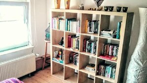 Une pièce avec une bibliothèque en bois remplie de livres, un radiateur, une fenêtre et un lit recouvert d’une couverture à rayures.
