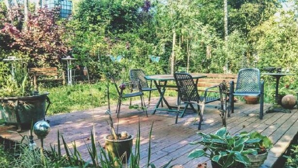 Un espace extérieur aménagé en terrasse, avec un sol en bois, des chaises et des tables en métal, le tout entouré d’une végétation luxuriante et de plantes en pot.