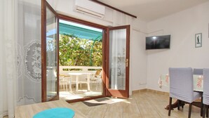 Ein Zimmer mit einem Holztisch, einem an der Wand montierten Fernseher und einer Glastür, die zu einem Außenbereich mit Tisch und Stühlen führt.
