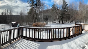 A snow-covered deck with a wooden railing, overlooking a forested area with trees and a small cabin. - Walk to the slopes from this BMOM 3 bedroom. (Rumford)