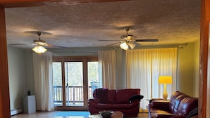 A spacious living room with a wooden floor, a red leather sofa, a matching armchair, and a coffee table. There are two ceiling fans and a large window with curtains. - Walk to the slopes from this BMOM 3 bedroom. (Rumford)
