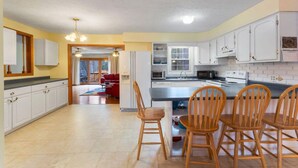 A kitchen with white cabinets, a central island with chairs, and a dining area in the background. - Walk to the slopes from this BMOM 3 bedroom. (Rumford)