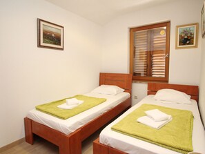 Habitación con dos camas de madera, cada una con ropa de cama blanca y mantas de adorno verdes. Dos cuadros enmarcados adornan las paredes, uno con un paisaje y el otro con un arreglo floral. Se ve una ventana con persianas de madera, y la habitación tiene un diseño sencillo y limpio.