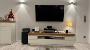 Ein modernes Wohnzimmer mit Flachbildfernseher an der Wand, einer Holzunterhaltungsecke darunter und einem Glastisch in der Mitte.