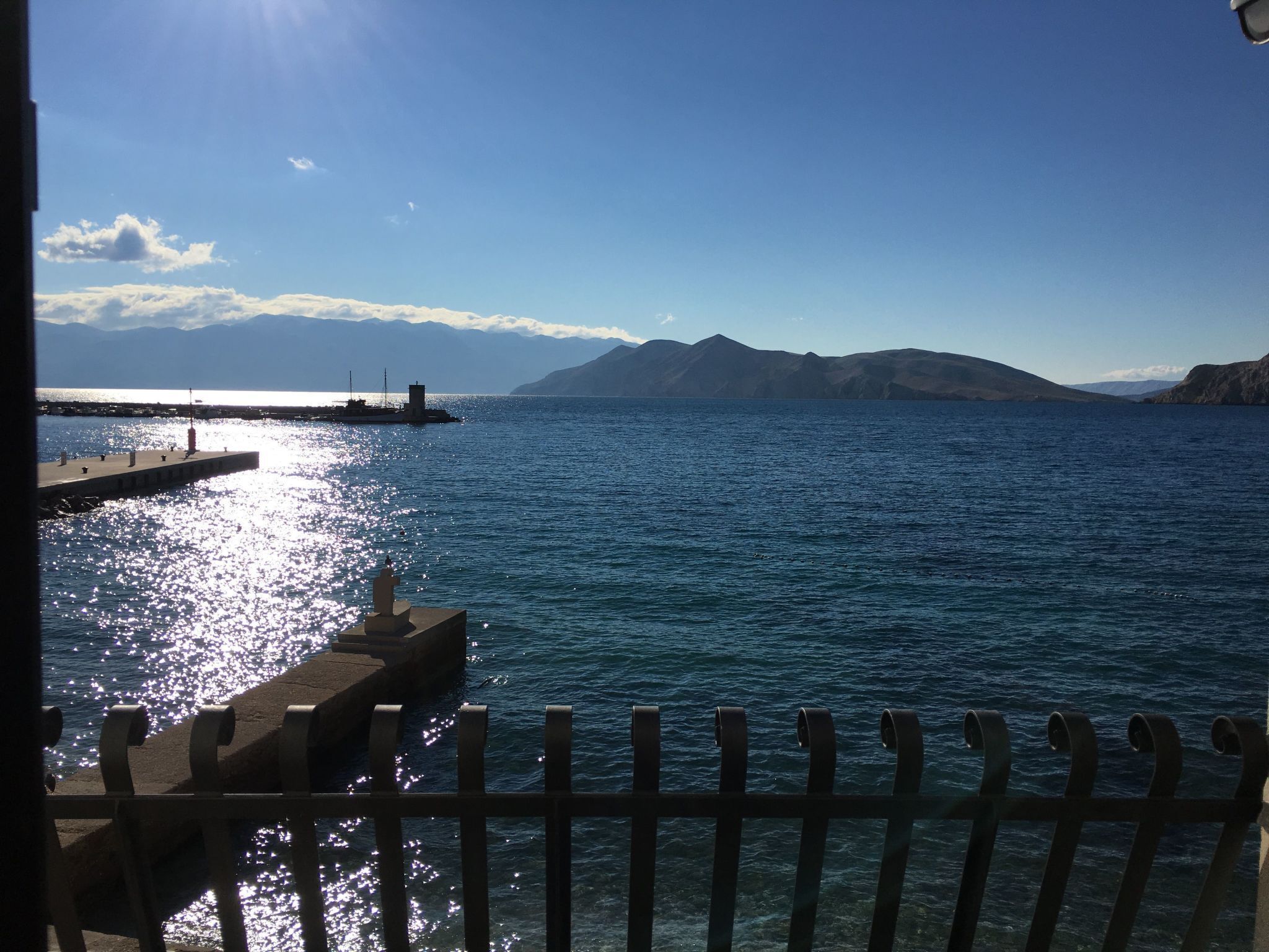 A coastal view with a pier extending into the sea, a clear sky, and distant mountains.