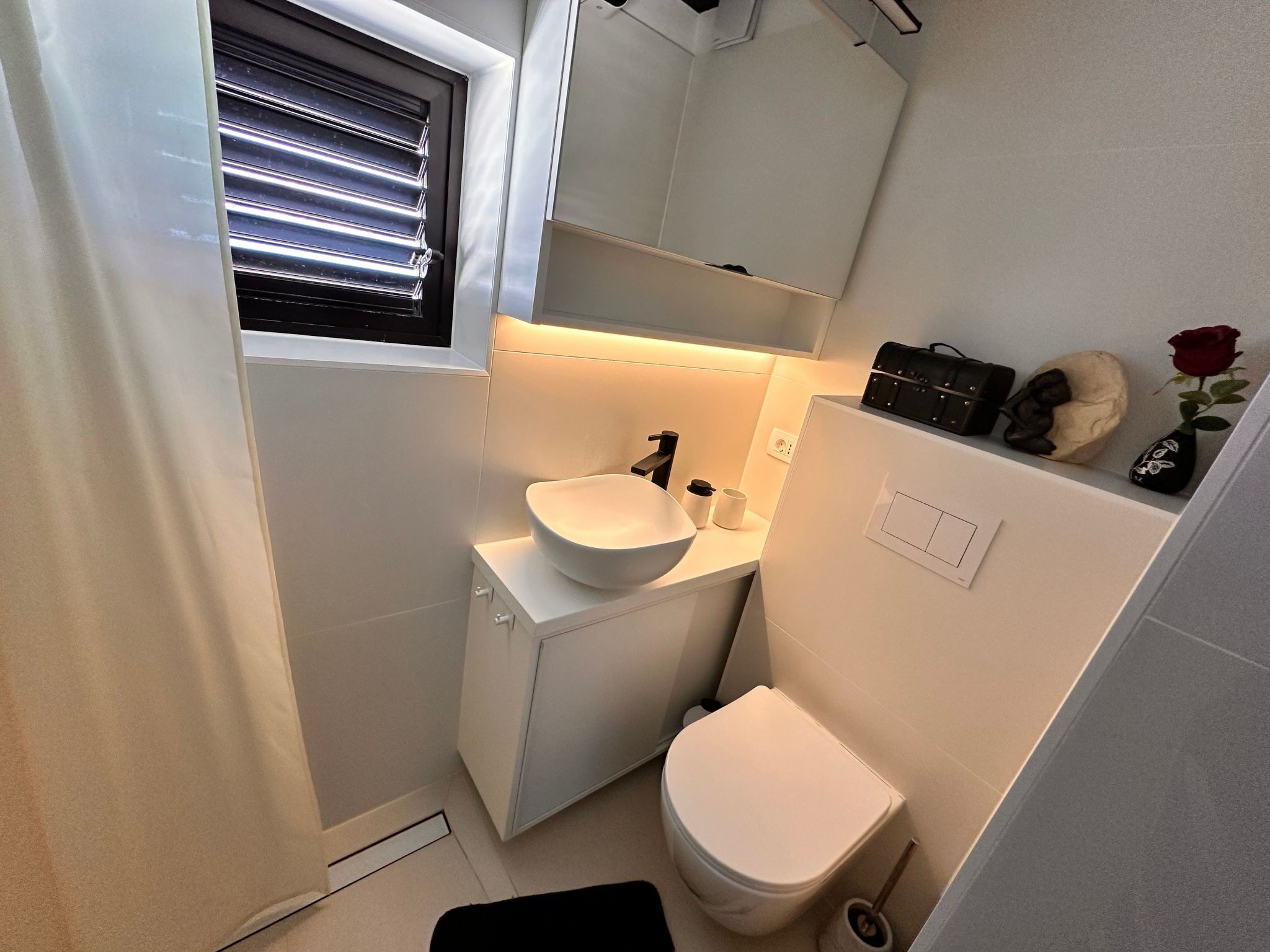 Un baño pequeño con lavamanos, inodoro y una ventana pequeña con persianas.