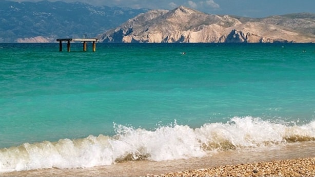 Une plage bordée d’une eau turquoise et limpide, des vagues qui se brisent sur le rivage et, en arrière-plan, une chaîne de montagnes rocheuses.