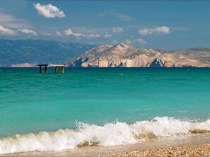 Ein Strand mit klarem türkisblauem Wasser, Wellen, die an die Küste schlagen, und einem felsigen Gebirge im Hintergrund.