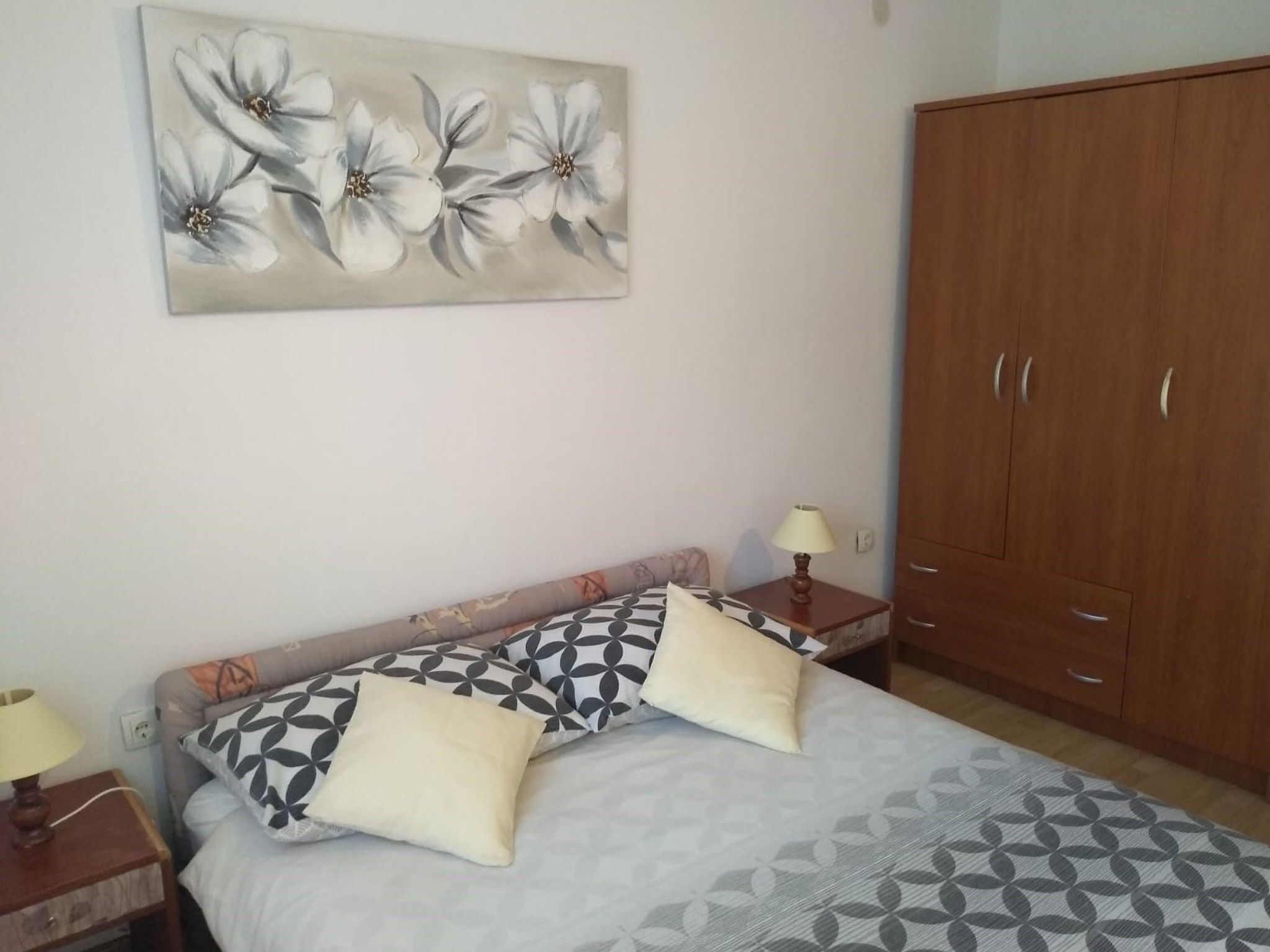 Une chambre à coucher avec un lit, deux tables de chevet avec des lampes, une armoire et un tableau représentant des motifs floraux accroché au mur.