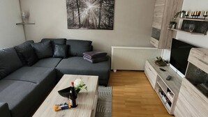 Ein modernes Wohnzimmer mit einem Ecksofa, einem Couchtisch mit Flasche und Blumen, einem Flachbildfernseher und einem Kamin mit Dekorationselementen.