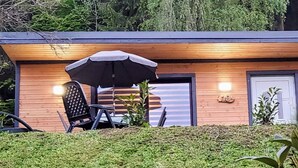 Ein modernes Gebäude mit Holzfassade, einer Tür mit Nummernschild und einer Terrassenfläche mit Tisch und Stühlen unter einem Sonnenschirm.