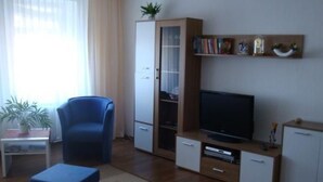 Un salon avec un fauteuil bleu, un meuble en bois sur lequel est posée une télévision, et une petite table sur laquelle se trouve un vase.