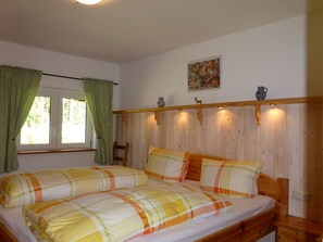 A bedroom with a wooden headboard, two beds with plaid bedding, a window with green curtains, a chair, a decorative shelf with small items, and a painting on the wall. - Irenahaus by Interhome (Auerbach)