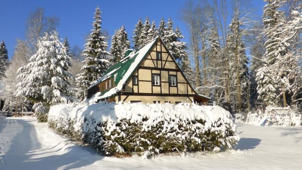 Un chalet traditionnel, doté d’un toit à forte pente, entouré d’arbres couverts de neige et surplombé d’un ciel bleu clair.