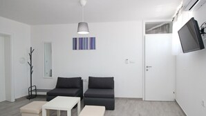 A modern living room with two sofas, a white coffee table, and a small ottoman. There is a ceiling light, a wall-mounted TV, and a door with a mirror. - Wohnung in Baška mit Terrasse by Interhome (Krk/Baška)