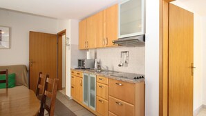 Cocina compacta con armarios de madera, microondas, estufa y lavamanos. Hay una mesa de comedor con sillas y un cojín verde.