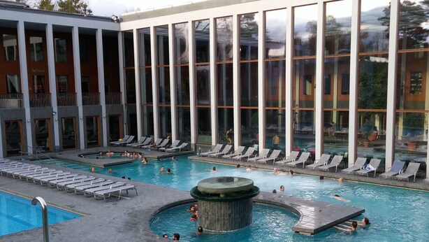 Una gran piscina coberta amb diverses línies de bades i una zona de banyera d'hidromassatge, envoltada de gandules i un edifici amb finestrals amplis.