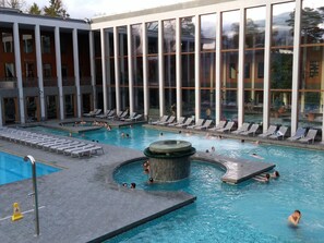 Una amplia piscina cubierta con varias pistas de natación y un área de hidromasaje, rodeada de sillones de descanso y un edificio con grandes ventanales.