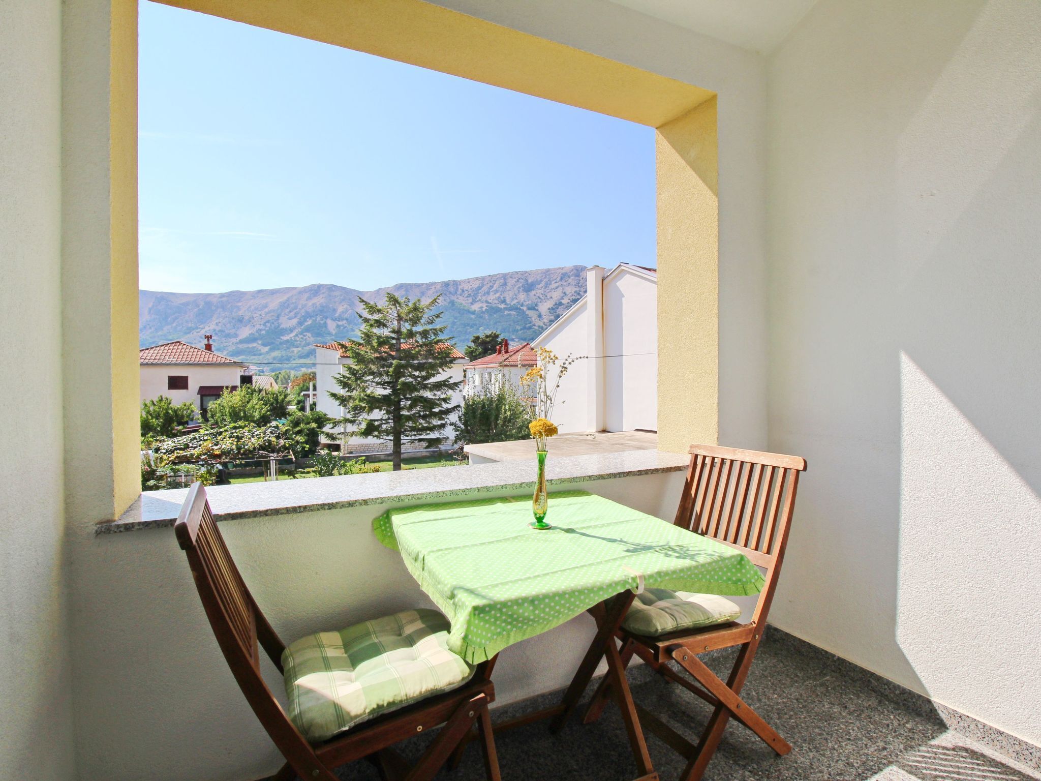 Ein Balkon mit Tisch und zwei Stühlen, einer Blumenvase und Blick auf Häuser und Berge.