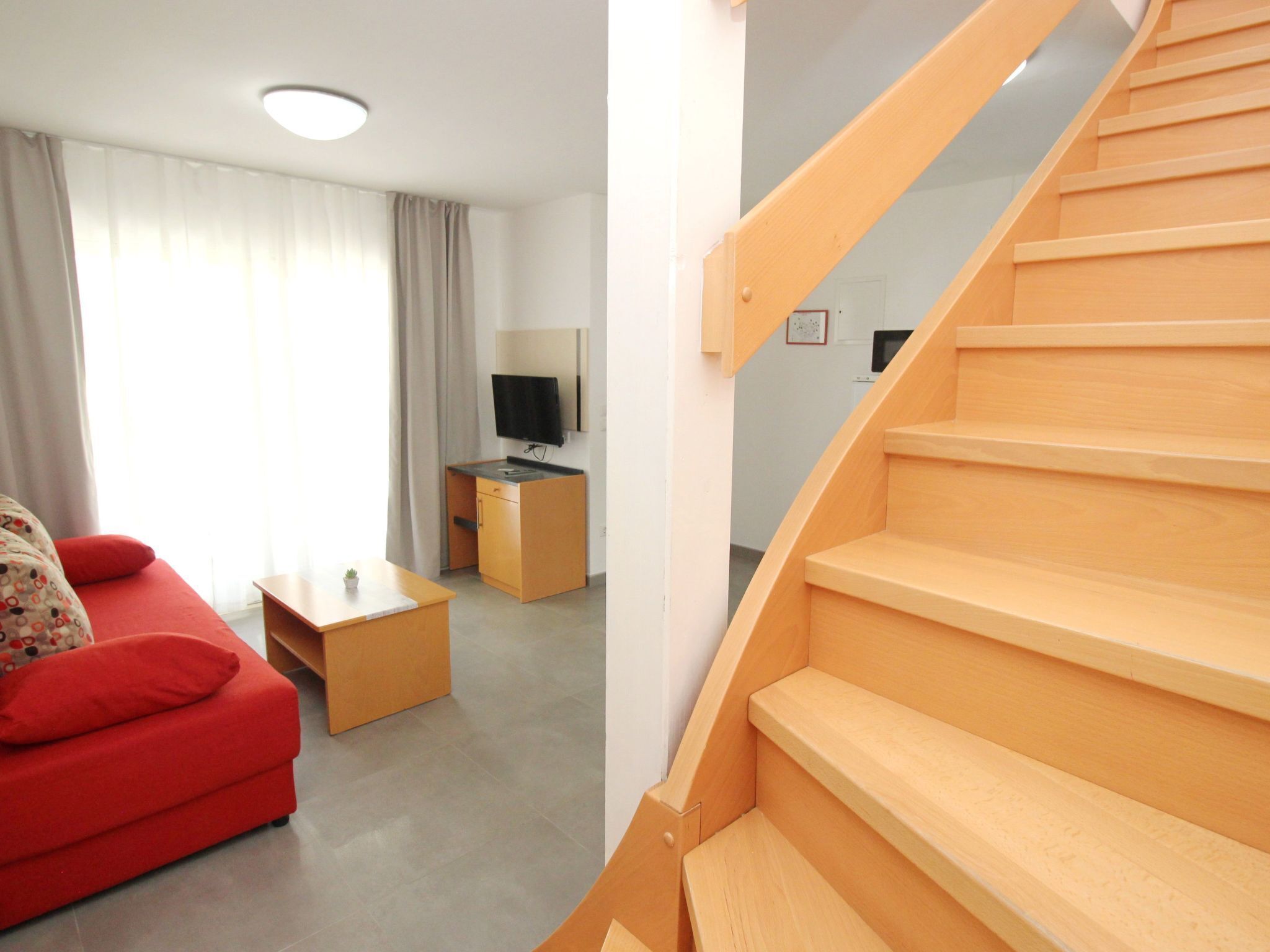 Una sala de estar moderna con un sofá rojo, una mesa de centro de madera, un televisor sobre un mueble bajo y una escalera de madera.