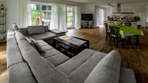 Een ruime woonkamer met een grote grijze hoekbank, een salontafel en een eethoek met een groene tafelkleed.