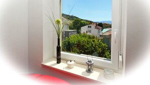 A window with a view of houses and greenery, a vase with plants, a teapot, and a lantern on a shelf. - Apartment Brnic8 by Interhome (Krk/Baška)