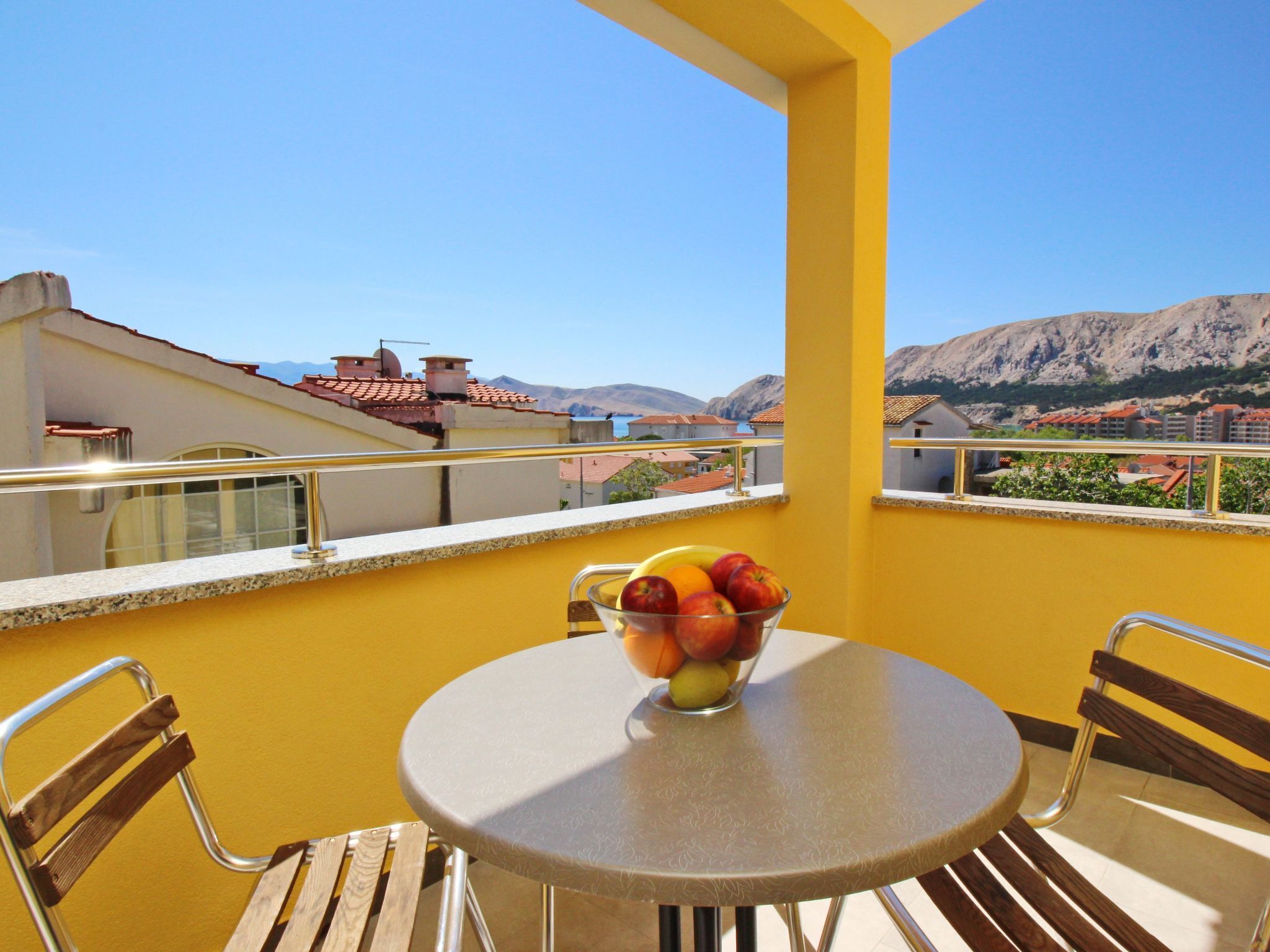 A balcony with a round table and chairs, a bowl of fruit, and a view of buildings and mountains.