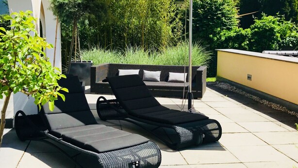 Eine Terrasse mit zwei Liegestühlen und einem großen Sonnenschirm, umgeben von Grünpflanzen und einer weißen Mauer.