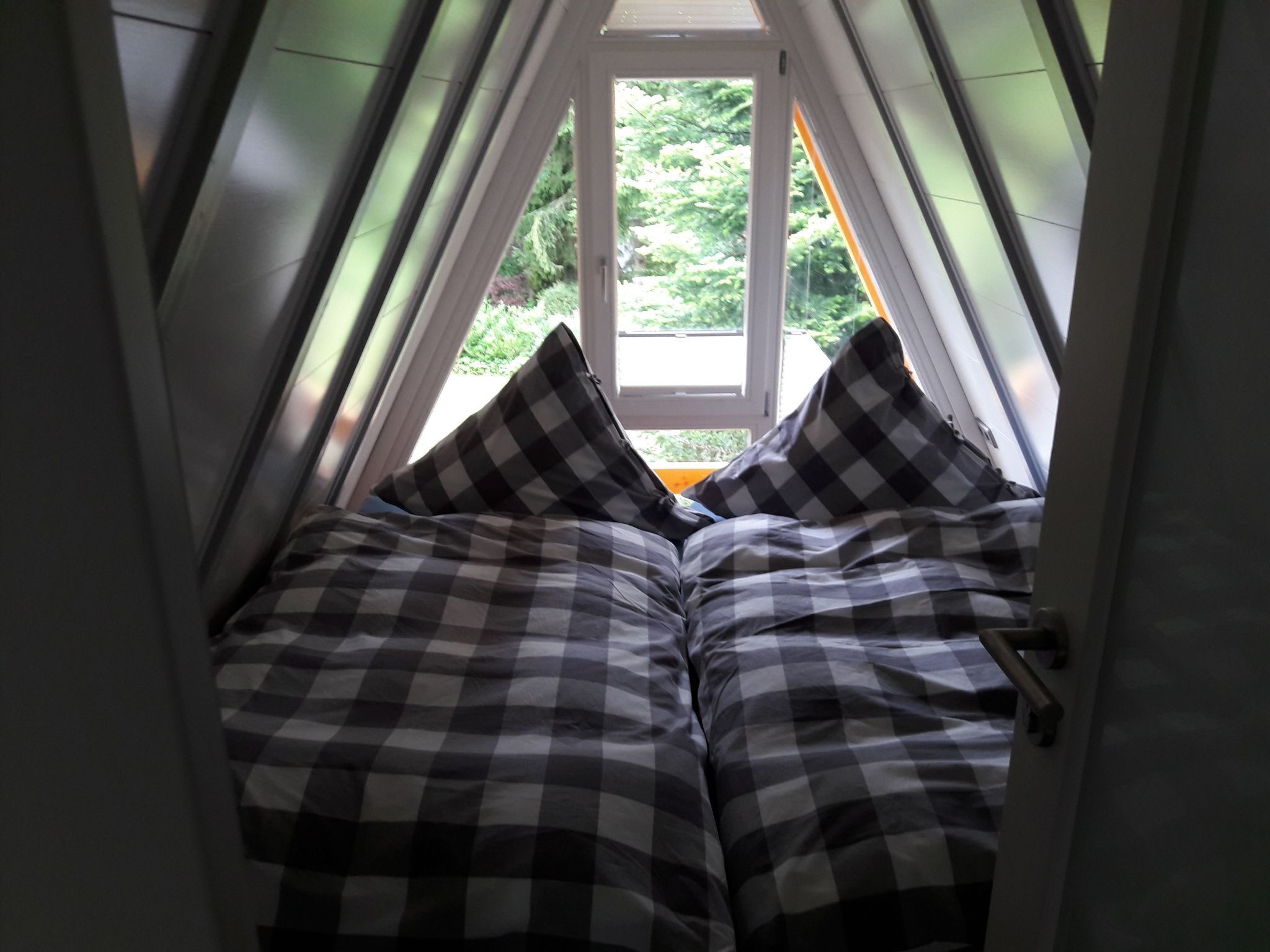 Ein schmaler, dreieckiger Raum mit einem Fenster am Ende, ausgestattet mit einem Bett mit kariertem Bettzeug und zwei Kissen.