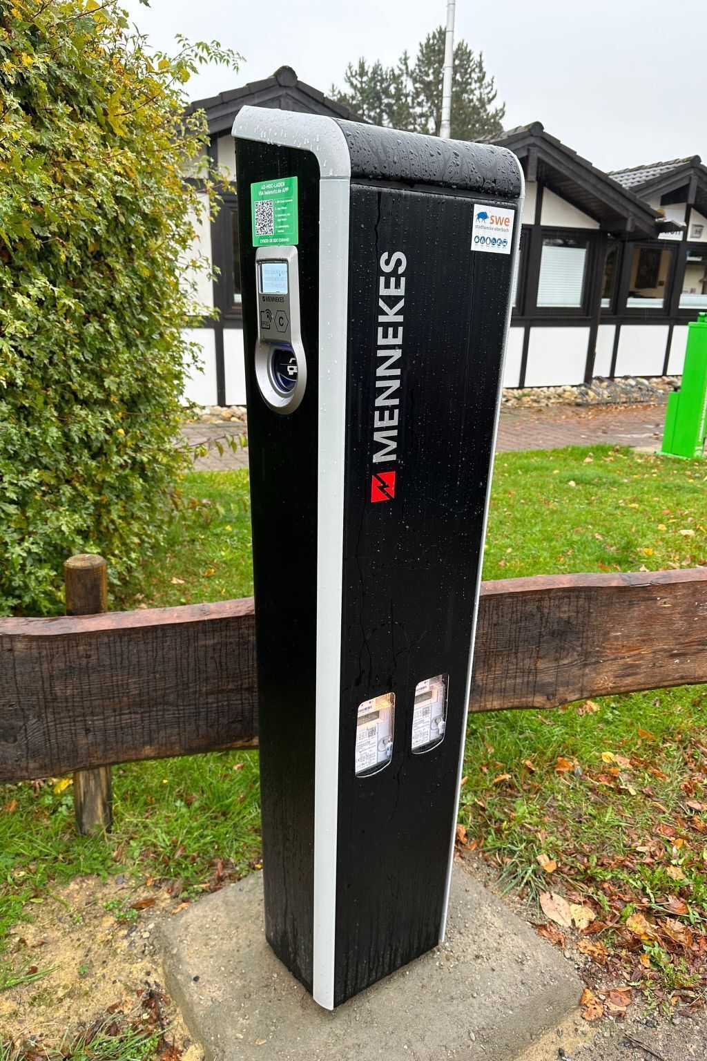 A black and white MENNEKES self-service kiosk with a QR code and a payment terminal, located outdoors with a wooden fence and greenery in the background.