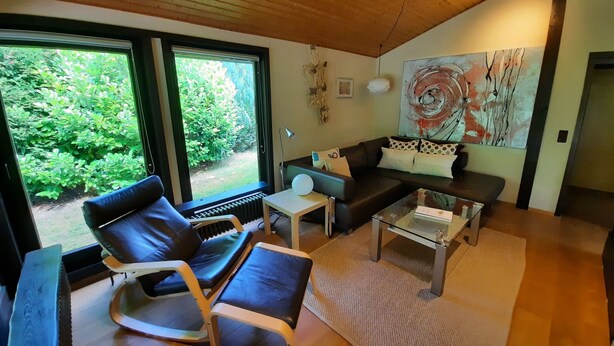 A modern living room with a black leather sofa, a glass coffee table, and a large window offering a view of greenery.